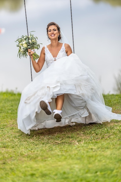 Heureuse mariée en robe de mariée blanche et bouquet de fleurs dans sa main se balance à l'extérieur à côté d'un lac