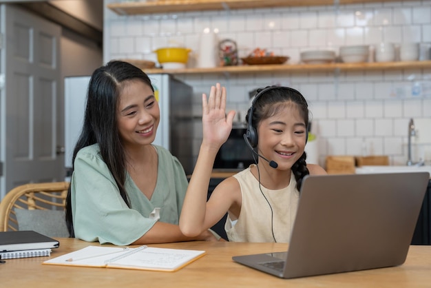 Heureuse mère asiatique et enfant assis à la table de la cuisine avec des crayons de couleur assistant à un cours de dessin virtuel par appel vidéo souriant et saluant l'écran de l'ordinateur portable pour saluer le professeur en ligne