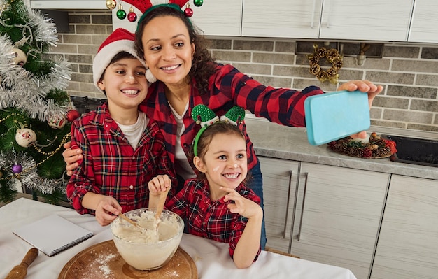 Heureuse mère en bonnet de Noel tient un smarphone dans les mains tendues et sourit en faisant un selfie serrant son beau fils et sa fille, préparant ensemble une pâte pour le pain et les biscuits de Noël