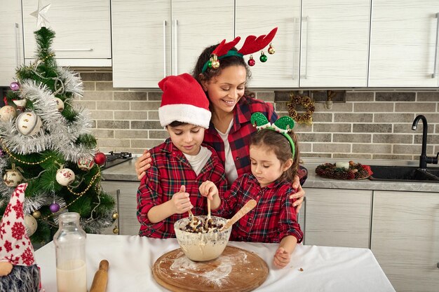 Heureuse mère embrassant son fils et sa fille, pétrissant une pâte pour préparer le pain de Noël. Ambiance de Noël, amour, tendresse, concept d'enfance heureuse et de maternité