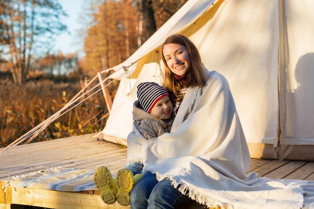 Heureuse mère embrasse son enfant avec une couverture tout en étant assis près d'une tente de camping