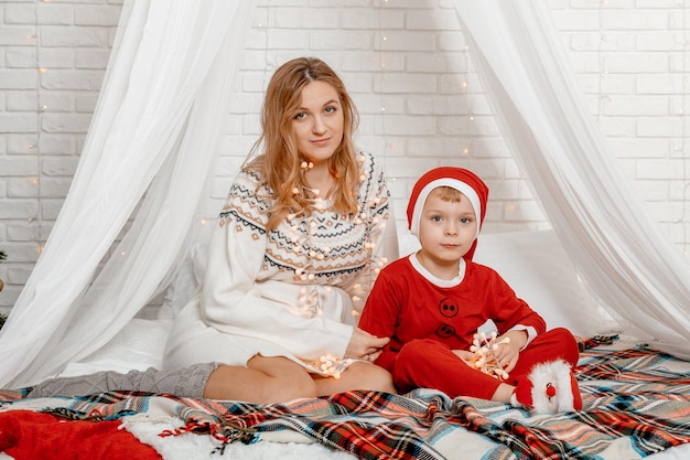 Heureuse mère enceinte et son fils regardant la caméra à Noël allongé sur un canapé sur le lit. Décor du nouvel an. La mère enceinte et son fils parlent et passent du temps ensemble au lit à la maison.