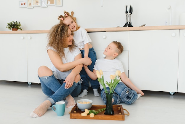 Heureuse mère étreignant sa fille et son fils assis sur un plancher en bois dans la cuisine à la maison.