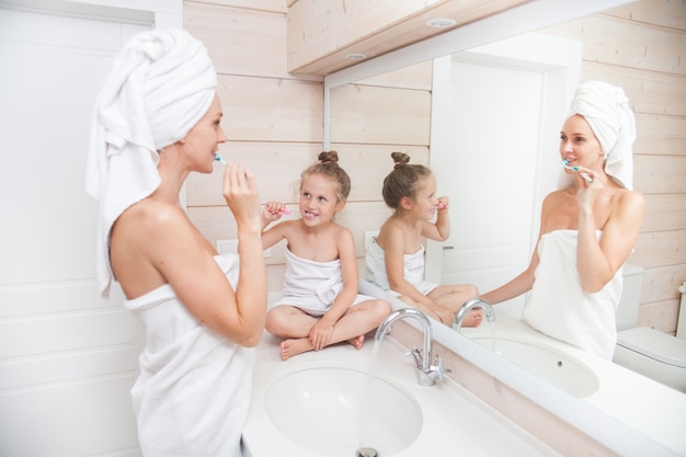 Heureuse mère de famille aimante et petite fille avec des serviettes se brosser les dents dans la salle de bain blanche.