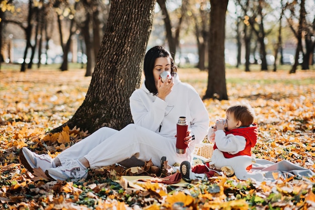 Heureuse mère de famille et petite fille en bas âge ayant un pique-nique d'automne avec thermos rouge et tasse