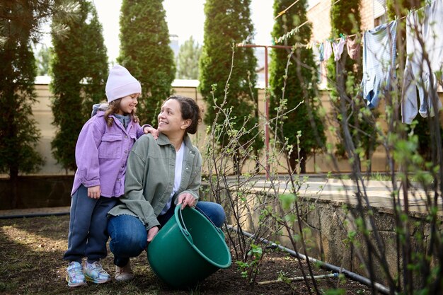 Heureuse mère et fille arrosant des buissons et des plantes à fleurs dans le jardin de l'arrière-cour d'une maison de campagne sur fond de séchage de vêtements sur une corde au début du printemps