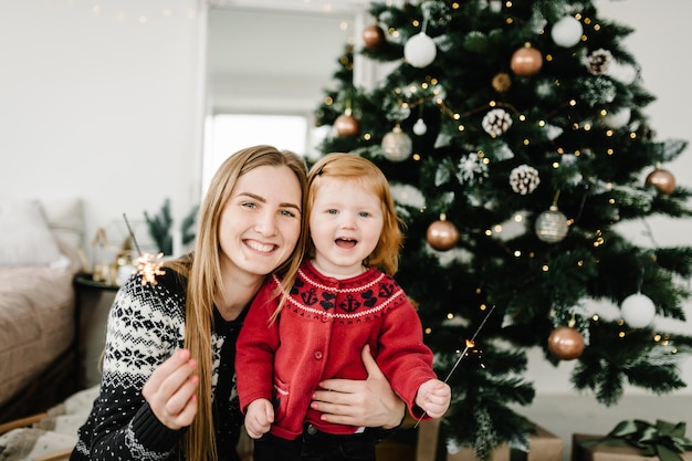 Heureuse mère et fille s'amusant avec des cierges magiques célébrant Noël à la maison Jeune maman et enfant tenant des cierges magiques étreignant et souriant près de l'arbre Joyeux Noël et bonnes vacances