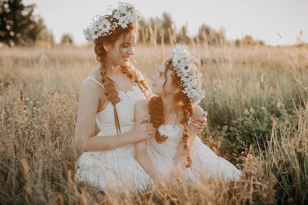 Heureuse mère et fille sourient et s'embrassant sur le terrain en été en robes blanches avec tresses et couronnes florales