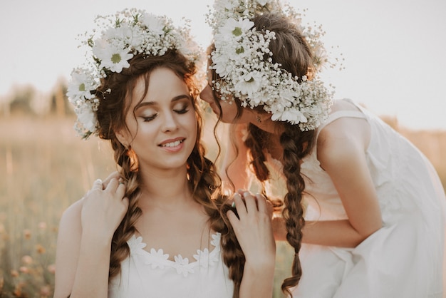 Heureuse mère et fille sourient et s'enlacent dans le domaine en été en robes blanches avec des tresses et des couronnes florales