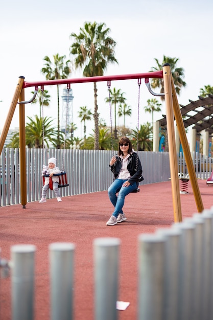 Heureuse Mère Et Jolie Petite Fille Se Balançant Sur Une Aire De Jeux Dans Le Parc D'été