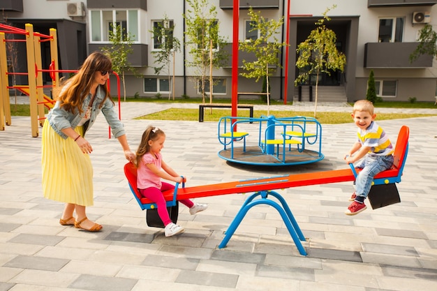 Heureuse mère jouant avec des enfants à l'extérieur. Enfants se balançant sur une aire de jeux moderne devant un immeuble