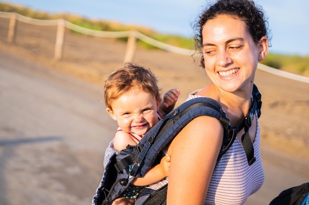 Heureuse mère portant un bébé souriant sur son dos dans un porte-bébé à l'extérieur