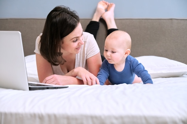 Heureuse mère souriante et son bébé allongés ensemble à la maison sur le lit et regardent l'ordinateur portable, le travail et les loisirs de la jeune mère