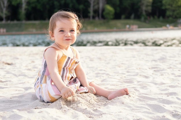 heureuse petite fille assise sur la plage sur le sable portant une robe d'été rayée aux beaux jours