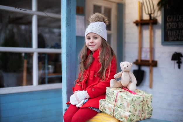 Heureuse petite fille assise sur le porche de la maison avec ours en peluche et cadeaux de Noël