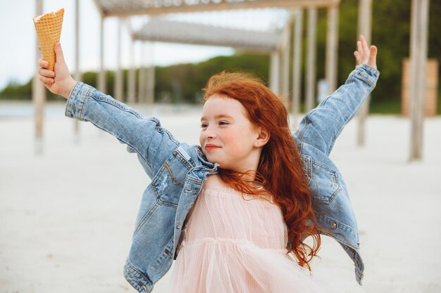 Heureuse petite fille aux cheveux rouges mange de la glace sur la plage en vacances d'été