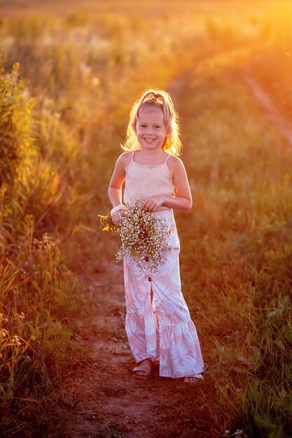 Heureuse petite fille caucasienne de 5 ans avec un bouquet de fleurs de camomille dans le pré en regardant la caméra, un portrait au coucher du soleil