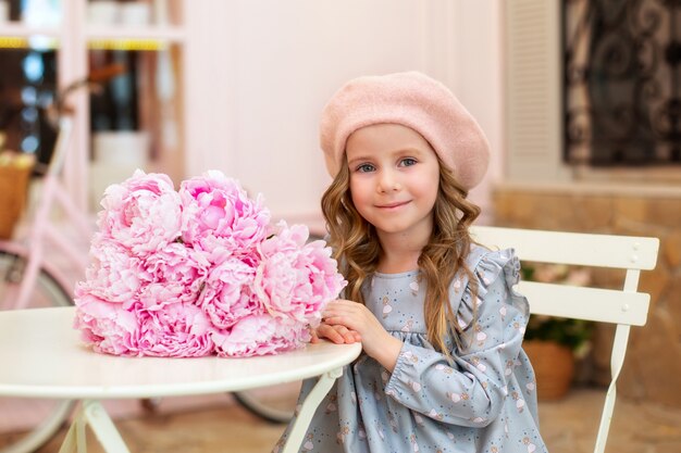 heureuse petite fille dans un béret est assise à table avec un bouquet de pivoines dans un café de style français