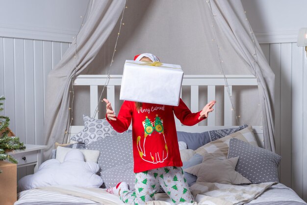 Heureuse petite fille drôle en pyjama et bonnet de Noel jette et attrape une boîte-cadeau au lit le matin de Noël