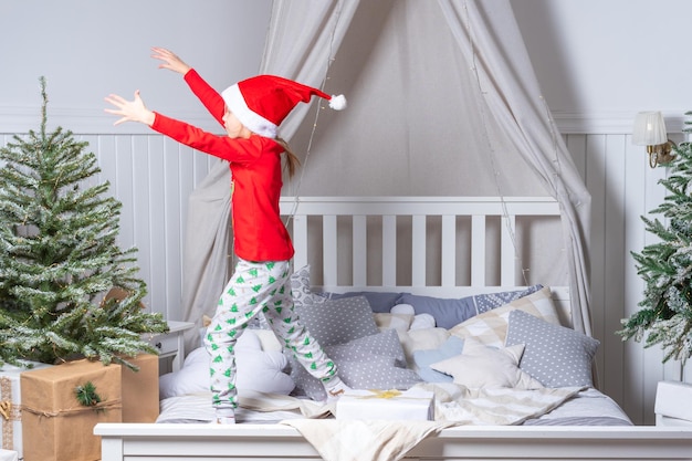Heureuse petite fille drôle en pyjama et bonnet de Noel saute dans son lit le matin de Noël L'enfant s'amuse