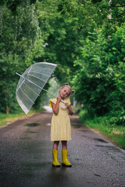 Heureuse petite fille émotive avec parapluie transparent en robe jaune et bottes de pluie dans le parc l'été