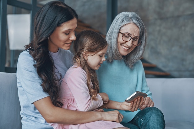 Heureuse petite fille examinant un téléphone intelligent tout en passant du temps avec sa mère et sa grand-mère à la maison