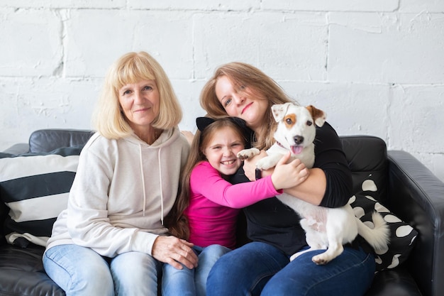 Heureuse petite-fille et grand-mère et fille assise sur un canapé avec un chien jack russell terrier. Grand-mère embrasse son petit-fils à la maison. Relation, famille et concept de trois générations.