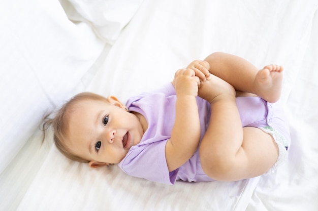 Heureuse petite fille mignonne dans un body est allongée sur le dos sur un lit sur des draps blancs, sourit, joue avec ses jambes. Le concept de produits pour bébés.