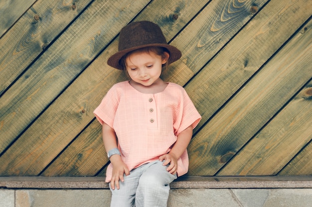 Photo heureuse petite fille mignonne dans un chapeau marron de style rural et des vêtements en mousseline dans une journée d'été sur zone en bois