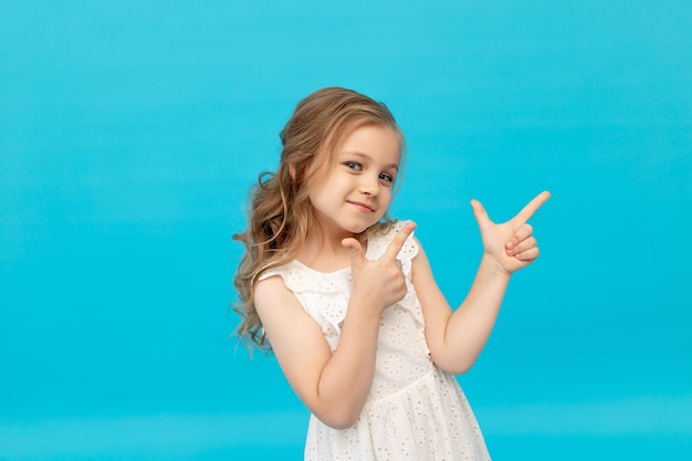 Heureuse petite fille mignonne dans une robe blanche en coton sur fond bleu dans le studio en riant en montrant ses doigts dans le ver, souriant et s'amusant, une place pour le texte
