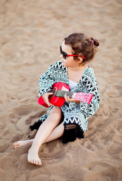 Heureuse petite fille mignonne jouant de la petite guitare ukulélé assis sur un lac ou une rivière à proximité de sable sur fond de nature