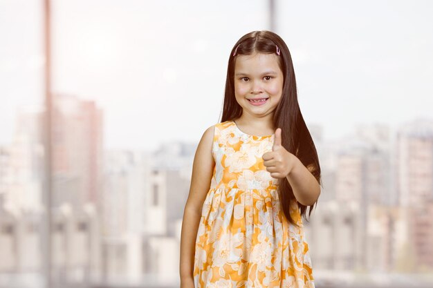 Heureuse petite fille mignonne montre le pouce vers le haut de l'arrière-plan flou des fenêtres intérieures