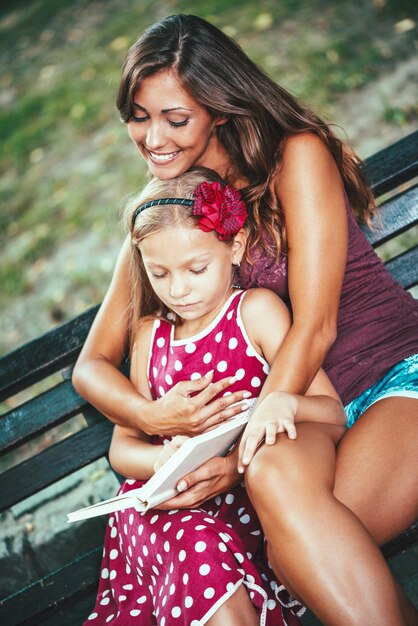 Photo heureuse petite fille mignonne et sa mère lisent un livre sur un banc dans le parc.