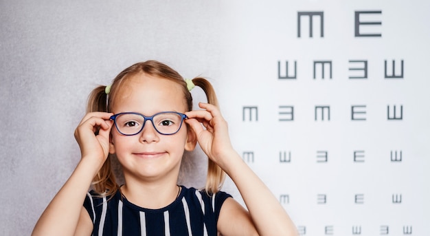 Heureuse petite fille portant des lunettes faisant un test de vue avant l'école avec un tableau des yeux flou à l'arrière-plan, la maternelle et l'examen médical scolaire