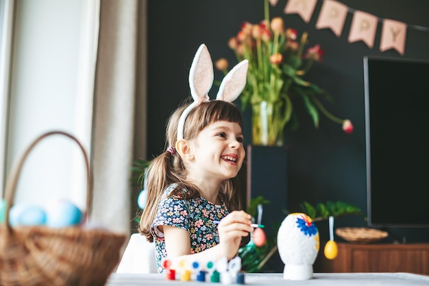 Heureuse petite fille riante avec des oreilles de lapin peignant des oeufs de Pâques en gypse