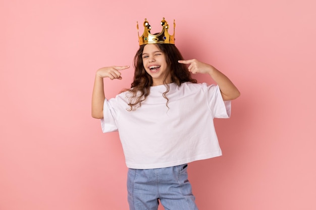 Heureuse petite fille souriante confiante debout dans une couronne d'or se pointant avec fierté