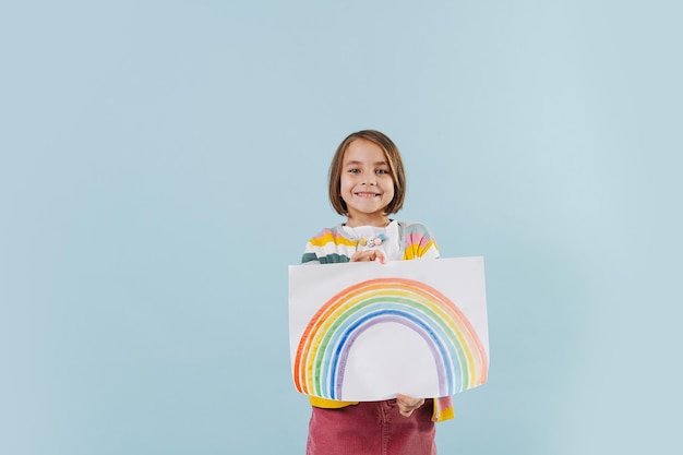 Heureuse petite fille tenant une peinture arc-en-ciel sur fond bleu