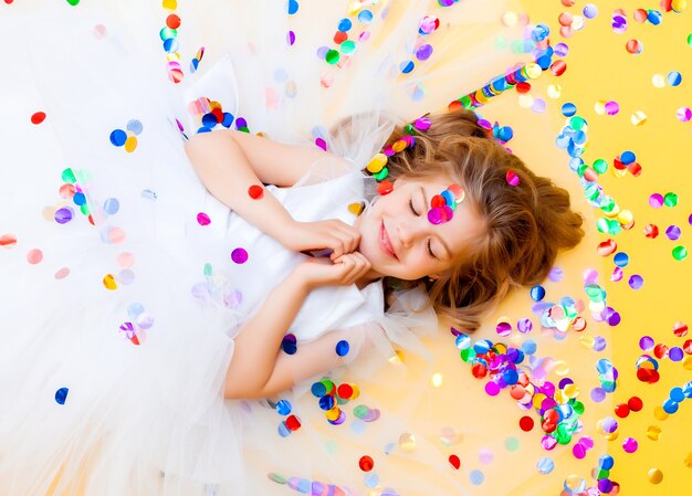 Heureuse petite fille vêtue d'une robe blanche célèbre une fête avec vue de dessus de confettis. Enfant d'anniversaire, émotions positives.