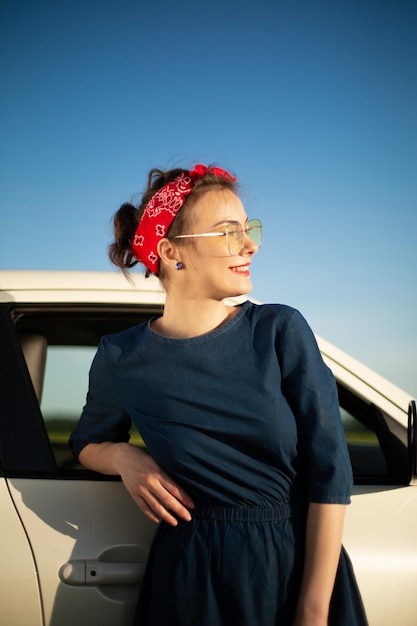 Heureuse positive souriante jeune jolie femme vêtue d'une robe bleue debout près d'une voiture blanche