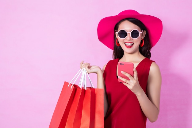 Heureuse robe asiatique jolie fille rouge tenant les sacs à provisions et téléphone intelligent à la recherche de suite sur fond rose