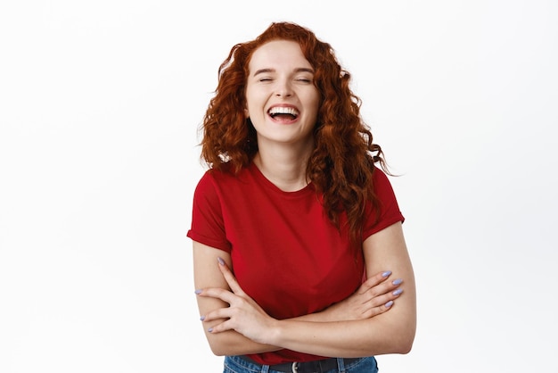 Heureuse rousse aux cheveux bouclés riant positivement les yeux fermés croisant les bras sur la poitrine et riant insouciant debout en t-shirt sur fond blanc