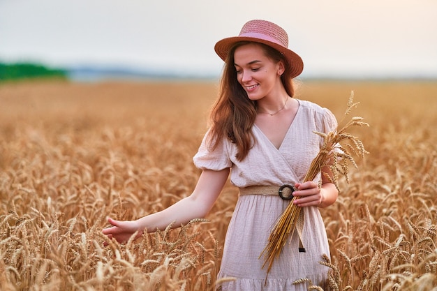 Heureuse souriante jolie jeune femme libre et mignonne portant un chapeau et une robe debout dans un champ de blé jaune doré et profitant d'une belle vie sereine en été