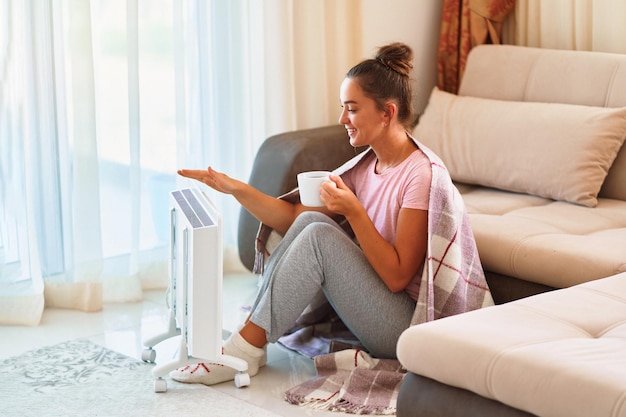 Heureuse souriante satisfaite joyeuse jeune femme avec des chaussettes à carreaux et tricotées buvant du thé chaud et se réchauffant près d'un radiateur électrique portable à la maison