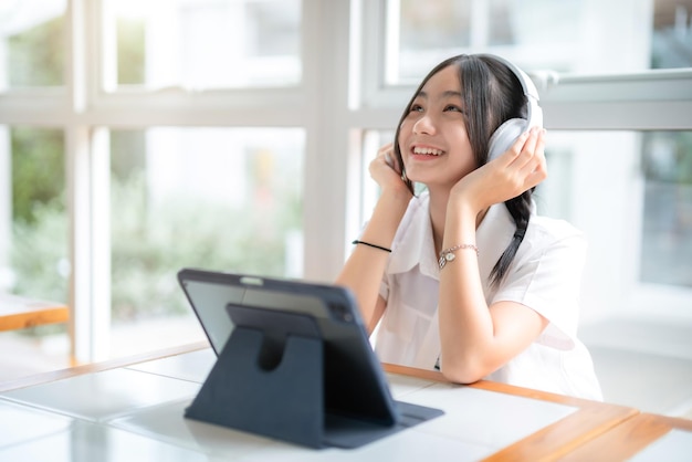 Photo heureuse de sourires mignons asiatiques de jeunes attirantes mignonne petite fille portant des écouteurs et écoutant de la musique en utilisant une tablette numérique travaillant à partir du caféconcept d'éducation et d'apprentissage en ligne