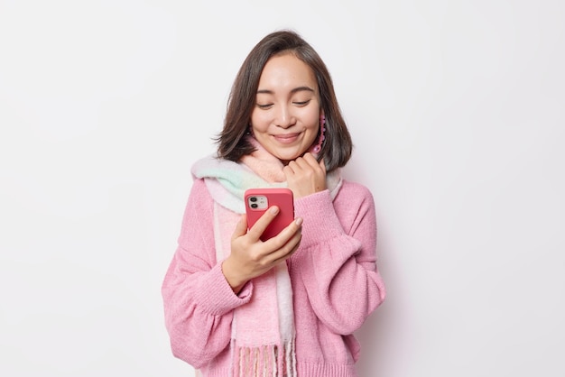 Heureuse tendre femme asiatique aux cheveux noirs regarde joyeusement le smartphone reçoit un message de son petit ami porte un pull chaud et une écharpe autour du cou isolé sur fond blanc regarde la diffusion