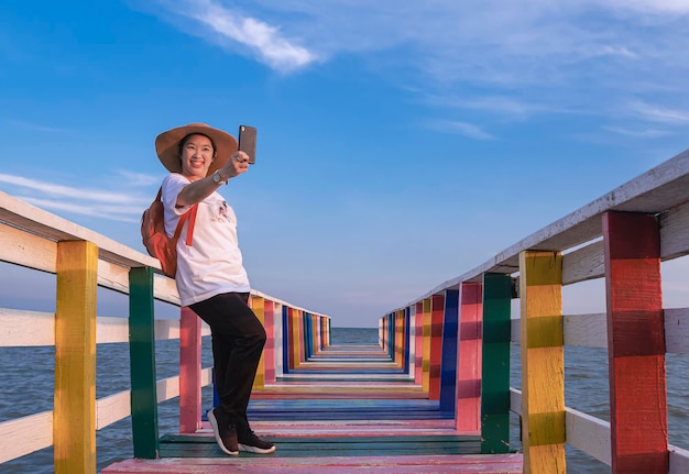 Heureuse touriste asiatique prenant un selfie avec un smartphone sur un pont en bois arc-en-ciel au point de vue de la mer