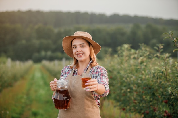 Heureuse travailleuse agricultrice souriante versant du jus de pomme savoureux dans un verre debout dans un verger pendant la récolte d'automne Temps de récolte