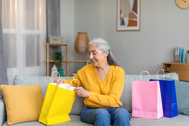 Heureuse vieille femme européenne aux cheveux gris regarde les achats dans le paquet après avoir fait ses courses dans le salon