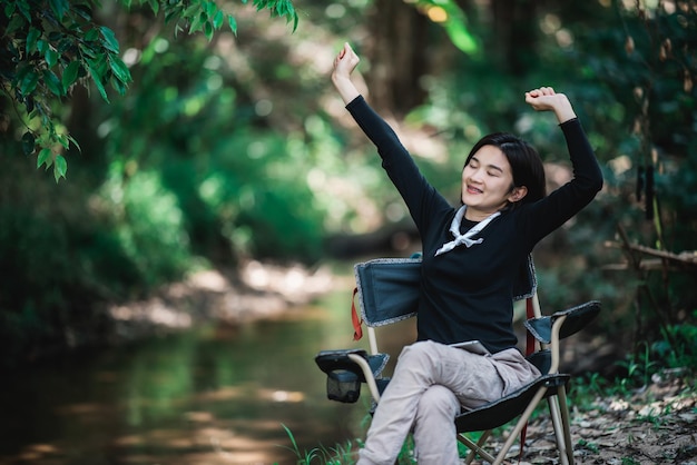 Heureusement jeune femme assise sur une chaise tout en se détendant sur le camping dans l'espace de copie de la forêt