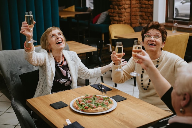 Heureuses femmes brunes et blondes vêtues de vestes noires et blanches, trinquant avec des verres de vin ou de champagne et riant de manière expressive au café célébrant la rencontre avec des amis et mangeant de la pizza